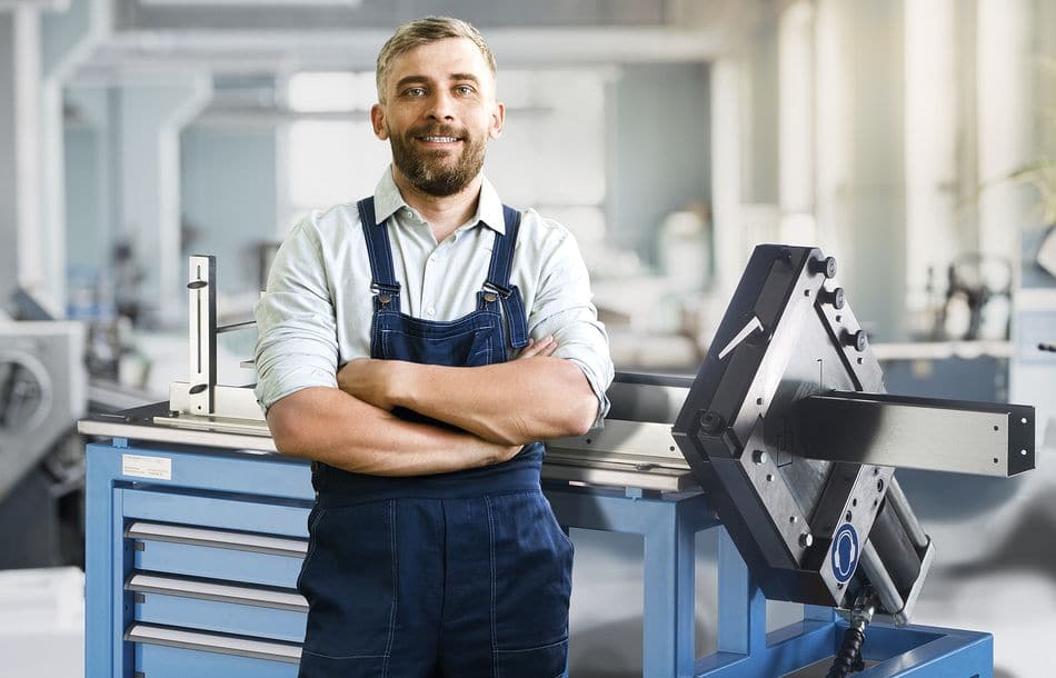 Cheerful content confident bearded typography worker in overall crossing arms on chest and looking at camera while standing among printing machines